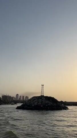 A remote rocky island stands in calm water. A metal tower sits atop the island. In the distance, a city skyline is visible, with construction cranes and smoke rising from a building. The sky is clear, with a gradient from light blue at the top to a warm yellow near the horizon, suggesting sunrise or sunset. The water appears choppy, with small waves. The overall scene is serene and hints at a coastal urban environment.
