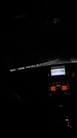 A car drives along a dark road at night. The only illumination comes from the car's headlights and the dashboard lights. The road appears to be winding, and there is sparse vegetation along the side. The car's radio is on, playing music. There are no visible people or interactions aside from the presumed driver.