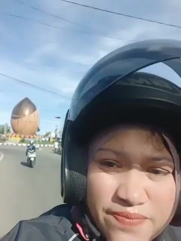 The video shows a first-person perspective of a person riding a motorcycle on a road. The rider is wearing a black helmet and a grey jacket. In the background, there is a large, brown, nut-shaped monument or sculpture on the left side of the road. Several other motorcycles and cars are visible on the road, along with traffic lights and power lines. The sky is blue with some clouds, indicating it is daytime. The overall scene appears to be a typical urban or semi-urban environment.