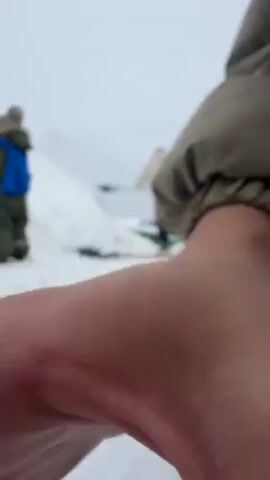 The video shows a blurry outdoor scene covered in snow, with three people present. In the foreground, the ground is blanketed with snow. To the left, a child in a dark green snowsuit with a blue accent is visible, standing and facing slightly away from the camera. In the center, an adult wearing pink pants, a khaki-colored jacket, and dark shoes is bent over, seemingly adjusting or working on something at their feet. In the background, there's a mound of snow and some bare trees on the right. The sky is overcast, creating a gray, muted atmosphere. The camera appears to be handheld, capturing a low-angle, slightly shaky view of the scene.