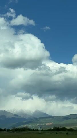 The video shows a landscape with mountains, clouds, and trees. The mountains in the background are covered in snow. The clouds are large and white against a blue sky. In the foreground, there is a field and some trees. The camera is moving, likely from a vehicle, as the scenery passes by. A street lamp is also briefly visible.