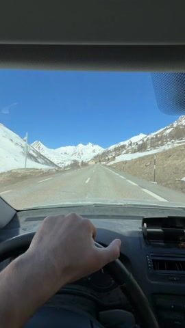 A first-person view from inside a car driving on a road through a mountainous area. The driver's hand is visible on the steering wheel. The landscape features snow-covered mountains under a clear blue sky. Another vehicle approaches from the opposite direction. Buildings can be seen along the roadside.