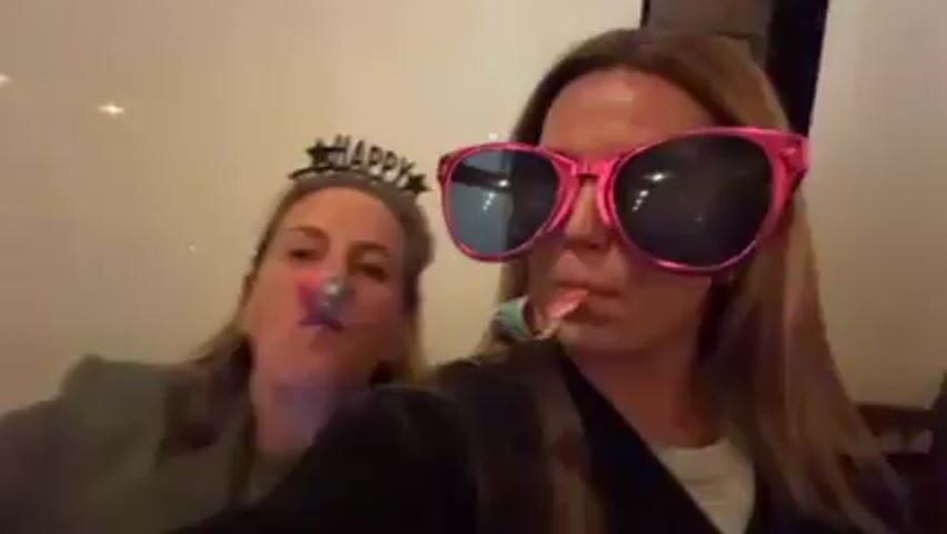 Two fair-skinned women celebrate New Year's. One wears a silver sequin Happy New Year crown. The other wears bright pink novelty sunglasses. Both hold party blowers in their mouths and blow. The camera focuses close on the woman wearing pink glasses who smiles. The camera tilts downward. A Happy New Year banner hangs above the heads of the two women. A wooden beam is visible on the wall behind them.