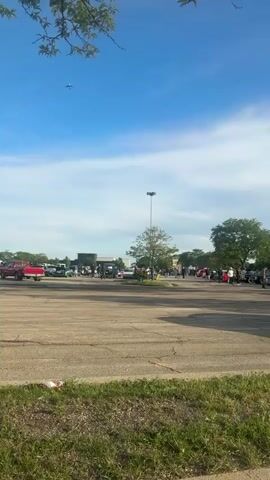 The video shows an outdoor gathering, likely a car show or meet, taking place in a large parking lot in Niles, United States of America. Numerous people are visible, walking around and congregating in groups, suggesting a social event. Many cars, including what appear to be classic or custom vehicles, are parked throughout the lot. A red pickup truck, a green car, and a white car are particularly noticeable among the diverse range of vehicles.

The setting includes several tall light poles, scattered trees, and buildings in the background. The sky is bright blue with scattered clouds, indicating a clear and sunny daytime. An airplane is briefly seen flying overhead at the beginning of the video. The atmosphere appears relaxed and engaging, with attendees socializing and observing the vehicles.