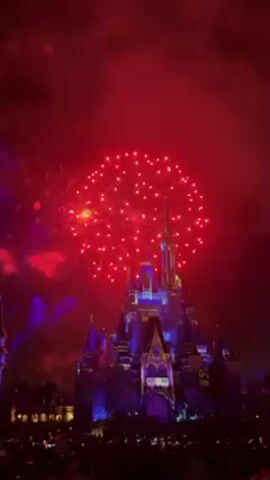 The video shows fireworks exploding over Cinderella's Castle at Disney World. The castle is illuminated in shades of blue and yellow. A crowd of people is visible in the foreground. The fireworks are mostly red, but there are also some white and yellow bursts. The sky is filled with smoke from the fireworks.