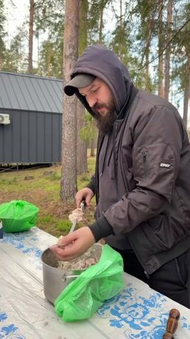 In an outdoor setting in Мишкино, Russia, a man is shown preparing meat for grilling. He is wearing a dark hooded jacket with "ZRD" visible on the sleeve, a dark hoodie underneath, and a baseball cap. He has a beard. The man is standing at a table covered with a white plastic tablecloth featuring blue floral patterns.

He is seen taking pieces of marinated meat from a metal pot and carefully threading them onto a metal skewer. The meat appears to be mixed with sliced onions and a creamy marinade. Next to the pot are two green plastic bags.

In the background, tall pine or fir trees suggest a forest or wooded area. A modern-looking dark grey building with a metal roof is also visible behind the trees, possibly a cabin or outbuilding. The lighting suggests it is daytime, likely overcast, given the attire and lack of harsh shadows. The man appears focused on the task.