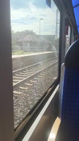 The video, captured from inside a moving train, shows a view through the window. Outside the window are train tracks running alongside a platform. A small, tiled-roof building is visible on the platform, along with a lamppost and some greenery. The sky is cloudy with patches of sunlight. The interior of the train is partially visible, with a blue and black patterned seat in the foreground. The train appears to be moving at a moderate pace, as evidenced by the slight blur of the passing scenery. There are no people visible outside the train, and the focus is on the landscape and the infrastructure of the train station. The lighting suggests it is daytime, possibly late morning or early afternoon. The overall impression is a serene journey through a rural or suburban area.