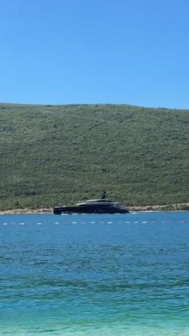 A black yacht is anchored in the blue ocean, with a green, tree-covered hill in the background. The water is clear and turquoise. There is a line of white and red buoys separating the yacht from the shore. The sky is clear and blue.