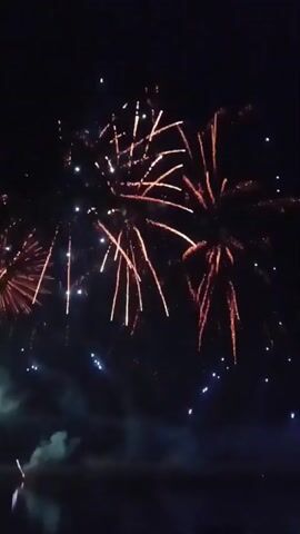 This video shows a fireworks display at night. There are several bursts of fireworks in various colors, including red, orange, and blue. The fireworks are illuminating the dark sky and reflecting on the surface of a body of water below. There is also some smoke visible in the lower left portion of the frame. The scene appears to be set at a waterfront location, likely a lake or river, with the dark water occupying the bottom of the frame. The fireworks are the central focus of the video, with the sky filled with their bright explosions.