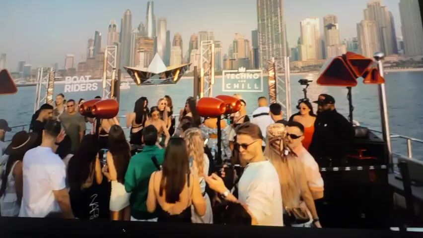 A group of people are gathered on a boat with a city skyline in the background. Some individuals are dancing, while others are standing and talking. There are two signs visible, one reading "BOAT RIDERS" and the other "TECHNO AND CHILL". The scene appears to be a party or event on the water.
