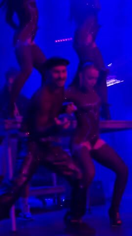 A man in a black sheer shirt, black beret, and black leather pants dances center frame. He has a mustache and is moving his hands. To his right, a woman in a black sequined bodysuit and thigh-high stockings dances. She is holding her left arm up. Behind them, other dancers in similar outfits are positioned on elevated platforms, with lights illuminating the scene in purple and red hues.