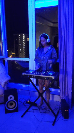 Woman DJs barefoot in blue-lit Sochi apartment overlooking cityscape