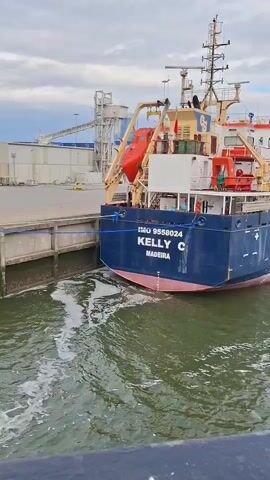 The video shows a vessel called "Kelly C" docked at a port. The ship's stern is prominently visible, displaying its name and IMO number. The water around the vessel is slightly disturbed, suggesting recent movement. In the background, there are industrial structures, including a tall conveyor system and what appears to be a wind farm in the distance. The sky is overcast, contributing to the muted lighting.