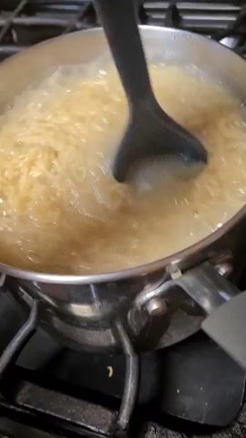 The video shows a close-up, slightly blurry, static shot of a pot of ramen noodles boiling on a stovetop. The noodles are a light yellow and appear to be fully submerged in the boiling water. A dark blue slotted spoon is partially submerged in the noodles, appearing to stir them. The pot is made of stainless steel and has two handles. The stovetop is black and appears to be made of metal grates. The lighting is somewhat dim. There is no audio.