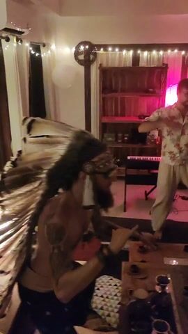 The video shows an indoor party with people dancing. The first shot features a person in a floral shirt and light pants standing in front of a keyboard. Behind them is a wooden shelving unit with lights. A coffee table is in the foreground. The video then shifts to show several people dancing enthusiastically. One man has his shirt off and is wearing loose, green pants. A woman in a black top and dark pants is also dancing. Other people are visible in the background, and the room has a casual, party-like atmosphere.