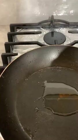 A person's hand is placing a ball of dough into a frying pan filled with oil. The dough is round and pale in color. The pan is black and appears to be heated, as evidenced by the sizzling oil around the dough. In the background, a gas stovetop is visible, with silver foil covering part of it. The lighting suggests an indoor setting, likely a kitchen.