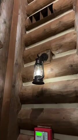 A close-up, low-angle shot shows a rustic lantern attached to a log wall. The lantern, made of dark metal and glass, has a bright bulb inside, casting a warm glow. The log walls have a rough, natural texture. Above the lantern, a wooden grate is visible. At the bottom of the frame, a small red box with a green display is partially visible. The camera pans slightly upward and to the right, focusing on the lantern and the surrounding log wall.