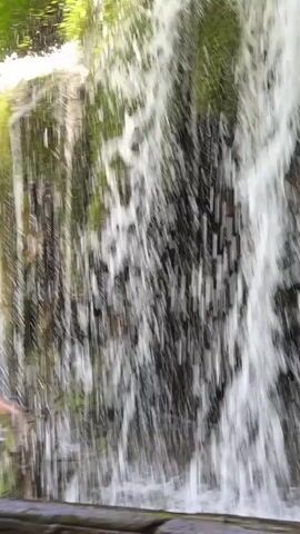 A video shows a waterfall cascading down a moss-covered rock face. The water is clear and foamy, with sunlight glinting off the droplets. Green foliage surrounds the waterfall, and a wooden railing is visible in the foreground. The scene suggests a natural, serene environment.