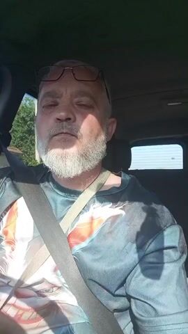 A middle-aged Caucasian man with graying hair and a beard, wearing glasses and a t-shirt with a colorful graphic, is sitting in the driver's seat of a vehicle. He is looking towards the camera and then up and to the left. The seatbelt is visible across his chest. The interior of the car is dark, and sunlight is coming in from the side, illuminating the man's face and t-shirt. The background outside the vehicle is blurry, showing some trees and buildings.