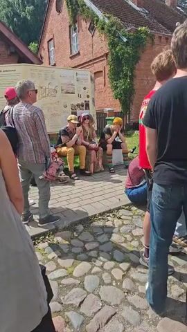 A group of people stand and sit outdoors in front of a red brick building with ivy growing on it. There is a large informational display board to the left of the people. Three individuals are seated on a bench in the center of the frame, two of them wearing colorful clothing and hats. The person on the left is a male wearing a patterned shirt and checkered pants. The person in the middle is a female wearing a patterned dress and a wide-brimmed hat. The person on the right is partially obscured but appears to be wearing brightly colored socks and shorts. Several other people are standing around them, some facing the seated individuals and others facing away. The ground is paved with cobblestones. The scene appears to be a public gathering or event.