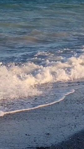 The video shows a close-up view of waves crashing on a beach. The water is blue and choppy, with white foam on the crests of the waves. The waves are breaking and rolling onto a dark, pebbly shore. The light suggests it is either early morning or late afternoon, casting a warm glow on the scene. There are no people or other objects visible in the frame. The sound of the waves is also captured.