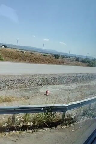 A video records a wide shot of a rural landscape under a clear blue sky. A paved road runs horizontally across the frame, with a gravel shoulder on the near side and dry grass and scattered bushes on the far side. In the distance, several utility poles and faint structures are visible, along with a large hill. A traffic cone lies on its side on the gravel shoulder in the lower left of the frame. The camera appears to be moving, possibly from inside a vehicle, as evidenced by the slight vibration and a partial railing visible at the bottom of the frame.