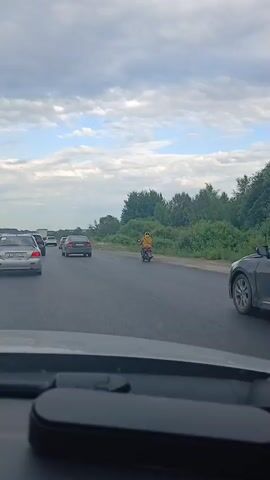 The media file captures a daytime scene on a highway in Белоярская Застава, Russia. The weather appears cloudy with patches of blue sky. The highway is moderately busy with cars traveling in the same direction. In the foreground, the dashboard of a vehicle is visible, suggesting the footage is from a car's perspective.

The primary subject of interest is a person on a scooter or small motorcycle, wearing a bright yellow jacket. This individual is traveling in the same lane as the cars, ahead of the viewer's vehicle. The scooter rider appears to be either cautiously navigating the traffic or possibly experiencing an issue. The presence of a scooter on a highway is a notable detail, as these are typically not designed for high-speed road use.

Several cars are visible on the road. To the left, a silver car with a license plate displaying Cyrillic characters is in front of the viewer. Further ahead, there are more cars in the distance. A dark-colored car is passing on the right side of the frame. The background consists of a line of trees and vegetation, typical of a roadside environment. The overall mood seems neutral, with the focus on the movement of traffic and the unusual sight of the scooter rider. There is no explicit indication of emotion, but the situation might evoke a sense of caution or concern due to the safety implications of a scooter on a highway.