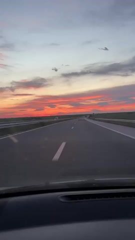 The video shows a view from inside a car driving on a highway at sunset. The sky is a mix of orange, pink, and grey clouds. The road is clear with white lines marking the lanes. There are sparse vehicles in the distance. The car's dashboard is visible at the bottom of the frame.