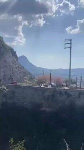 The video shows a scenic landscape featuring a mountain range and a road elevated by a stone wall.  The road has occasional car traffic and a few people are standing on the shoulder. The sky is blue with scattered clouds, and sunlight shines through. Tall power poles line the road. Below the road, there is foliage.