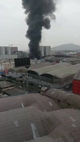 The media file depicts a dramatic industrial fire in Breña, Peru. A massive plume of thick, black smoke billows into the sky, dominating the upper portion of the frame. The smoke rises from a low-lying structure, likely a factory or warehouse, situated amidst a dense urban landscape.

In the foreground, the roof of a corrugated metal building is visible, suggesting the vantage point is from a nearby elevated structure. Beyond this, a large industrial building with a curved, light-colored roof stretches across the mid-ground. A street runs along the side of this building, with a few vehicles, including cars and what appears to be a truck, visible on it.

The background is characterized by a cluster of modern, multi-story buildings, some of which are tall apartment complexes. The architecture suggests a developed urban area. The overall scene is one of urban industrial activity juxtaposed with the destructive force of a fire.

The weather appears overcast, with a hazy, diffused light, suggesting it could be morning or late afternoon. There are no discernible people in the immediate vicinity of the fire, though the presence of vehicles on the street implies general urban activity. The overwhelming visual element is the intense smoke, creating a sense of urgency and potential danger.

There is no visible text in the media file. The emotions evoked are primarily concern and alarm due to the nature of the event. The location is identified as Breña, Peru, which provides the geographical context for this urban industrial fire.