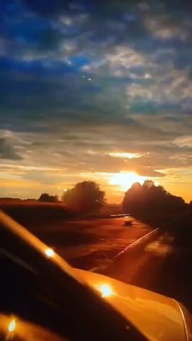 The video shows a view from inside a car looking out at a road during sunset. The sun is setting on the horizon, casting a warm orange glow across the sky and reflecting off the road and the car's hood. The sky is filled with dramatic clouds, ranging from deep blues to soft oranges and yellows. Trees and fields line the side of the road, silhouetted against the bright sky. The camera appears to be mounted on the dashboard or windshield. The car is in motion, indicated by the blurred scenery and the changing perspective. There are no people visible in the frame.