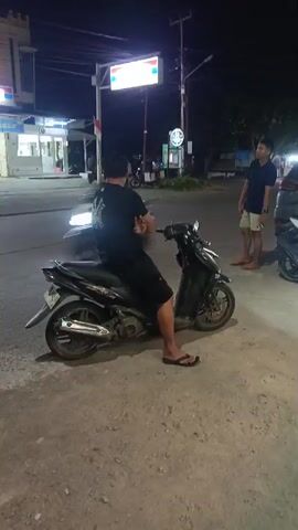 The video begins at night on a street, possibly near a gas station or convenience store. In the foreground, a person is sitting on a motorcycle that is in motion, indicated by motion blur. Further down the street, a person stands near a parked car. The scene then transitions to a wider view of the street at night, with cars and streetlights visible. The moon is also visible in the dark sky. Two young people are standing near a scooter, looking upwards. The video then shifts to an indoor setting, possibly a food stall or small restaurant. People are seen behind a counter and customers are waiting. A sign with green lettering is visible above the counter. The video concludes with a clearer view of the indoor scene, showing people interacting and ordering food.