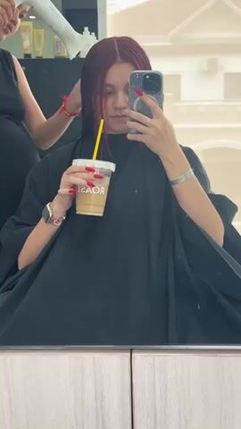 A person with long, dark red hair, wearing a black cape, is holding a phone and a clear cup with a beverage and a yellow straw. They are taking a selfie in a mirror. Another person, partially visible on the left, is holding a white hairdryer and appears to be drying the first person's hair. The environment looks like a salon, with shelves and bottles visible in the background.