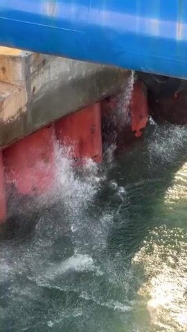 The video shows a close-up view of the underside of a large blue structure, possibly a ship or a bridge, as it moves through choppy green water. Several large, red cylindrical supports are visible, extending downwards from the blue structure and disappearing into the water. The water is splashing and churning around these supports, indicating movement and possibly some resistance. The lighting suggests it is daytime, with sunlight reflecting off the water's surface. There are no people or animals visible in the frame. The focus is on the interaction between the structure and the water.