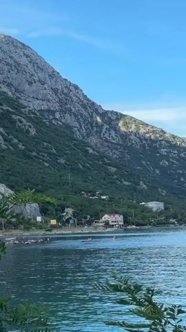 The video begins with a wide shot of a bay with a mountain range in the background. Several people are in the water, some appearing to be on paddleboards, and others swimming. A few buildings are visible along the shoreline, nestled at the base of the mountain. As the video progresses, the camera zooms in on the mountain. A small, arched opening, resembling a tunnel entrance, is visible high up on the rocky slope, partially obscured by green foliage. The sky is a clear blue.