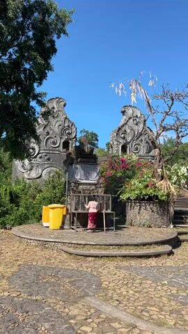 A person wearing a red and white patterned sarong and a light-colored top is standing at a table with a yellow trash can to their left. The person is holding an object, possibly a broom or a mop, and appears to be cleaning the area. Behind them is an ornate stone structure with intricate carvings and a checkered cloth hanging in the center. Lush green foliage and a dry, thorny tree frame the structure, and the sky above is a clear blue. The ground is paved with cobblestones, and there are stone steps leading up to the right of the scene.