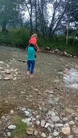 A person is carrying a child on their shoulders while walking across a shallow river. The person is wearing a teal shirt and blue pants, and the child is wearing a pink shirt. The riverbed is covered in rocks, and there is vegetation on the banks. The water is clear and appears to be flowing gently. The trees on the bank are tall and leafy, and the sky is overcast.