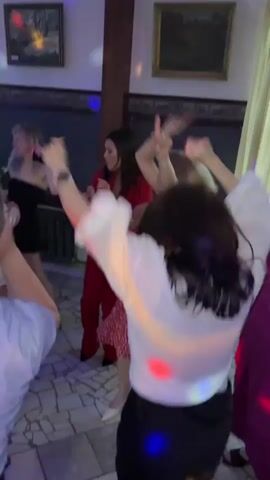 The video shows a group of people dancing in a room with a tiled floor and large windows. Several individuals are visible, mostly women dressed in party attire. One woman in the foreground is wearing a white top and black skirt. To her left, a woman in a pink outfit is dancing. In the center, a woman in a black strapless dress is visible, and to her right, a woman in a red jumpsuit is dancing. Further back, a woman in a red dress and another in beige pants and a dark top are also present. The room is dimly lit, with colored lights projecting onto the floor and walls. Two framed pictures hang on the wall. The atmosphere appears to be a celebration or party.