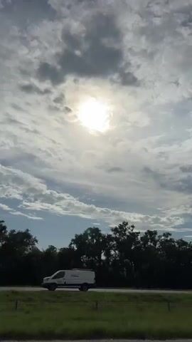 The video captures a view of a highway under a partly cloudy sky. The sun is visible and partially obscured by clouds, casting a bright glow. Trees line the side of the highway, and several cars are seen driving in both directions. The camera appears to be mounted on a moving vehicle, likely traveling on the highway itself, as evidenced by the consistent movement of the landscape. The foreground shows a guardrail and the edge of the road. The overall impression is of a typical daytime commute or travel scene.