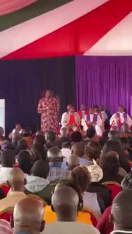 A woman in a long dress with a floral pattern is speaking from a stage in front of a large crowd. Behind her, sitting on chairs, are several men in clerical robes. The space is decorated with red, white, and purple drapes. The audience is comprised of men and women of varying ages. The speaker is holding a microphone. The atmosphere seems formal and respectful.