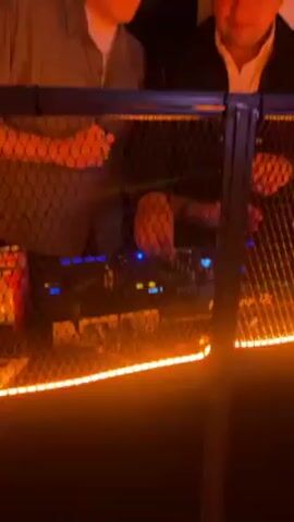 Two men stand behind a DJ booth with black mesh covering the front, and an orange light stripe running along the bottom. One man wears a grey shirt over a white undershirt and headphones. The second man wears a black Nike baseball cap and a black jacket over a white shirt. Behind them are two disco balls, a white speaker, candles in red holders, and green plants. The man with the headphones raises his right hand up to his ear. The man in the baseball cap looks down at the DJ equipment.