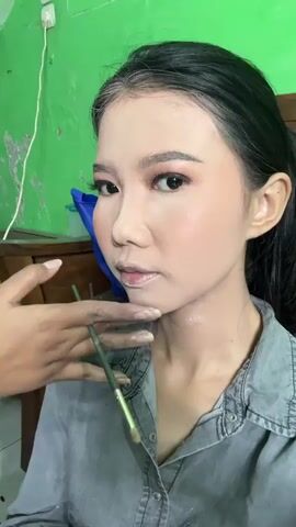 A medium shot shows a woman with dark hair and light skin, wearing makeup and a denim shirt, looking to the right of the camera. Her face is made up with blush, dark eyebrows, eyeliner, and pink eyeshadow. She has some white powder on her neck. A hand wearing a red shirt is holding a makeup brush near her chin. The background is a bright green wall and a wooden table.