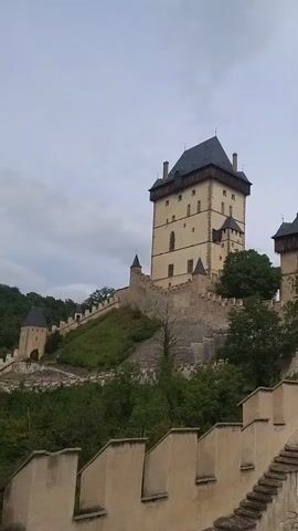 This video shows a panoramic view of a medieval castle complex under a cloudy sky. The castle, made of stone and light-colored plaster, features several towers with dark, pointed roofs. A prominent tall tower with multiple windows stands on the left, while other buildings with similar architectural styles are visible to its right. A long, fortified wall with crenellations runs along the foreground, leading to a set of stone stairs that ascend towards an arched entryway. Lush green vegetation, including trees and bushes, surrounds the castle and its walls, indicating a natural, outdoor setting. The camera pans slowly from left to right, offering a comprehensive view of the castle's exterior and its imposing fortifications.