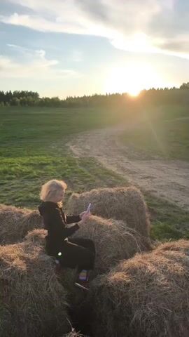 A young woman with curly blonde hair, dressed in a black tracksuit and colorful socks, is sitting on a stack of hay bales in a rural field. She is holding a smartphone with a light purple case and appears to be taking photos or videos, pointing the phone towards the horizon. The setting sun is visible in the background, casting a warm, golden glow across the landscape and causing a lens flare on the camera. The field has green grass and a dirt path leading into the distance, with a treeline bordering the horizon. The sky is partly cloudy with patches of blue. The overall scene suggests a tranquil evening in the countryside, possibly in Стеклянный, Russia, as the woman captures the sunset.