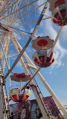 The video shows a Ferris wheel with several gondolas in motion against a blue sky with white clouds. The gondolas are red and white with decorative elements. One gondola is closer to the foreground, and another is further up and to the right. A ladder is visible on the side of the Ferris wheel structure in the lower left. The bottom portion of the Ferris wheel's structure, which appears to be part of a carnival ride, is visible, featuring decorative panels and lights. The Ferris wheel is slowly rotating, causing the gondolas to move.