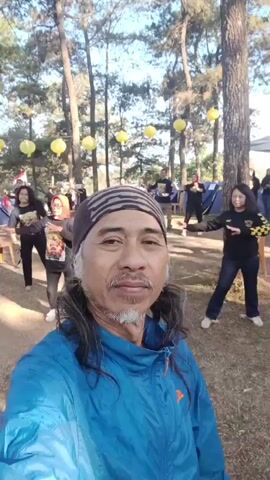 The video shows a man in a blue jacket and a bandana taking a selfie in an outdoor setting with trees and yellow lanterns. In the background, several people are seen engaged in what appears to be a group exercise or dance. One woman in the foreground on the left is wearing a red hijab and a black t-shirt with a graphic print, and she is in a dynamic pose. To her right, another woman in a black shirt with yellow accents is also in motion. Further back, more individuals are visible, some with their arms raised. Tents and chairs are scattered around the area, suggesting a communal gathering or event. The overall atmosphere seems to be lively and outdoorsy.
