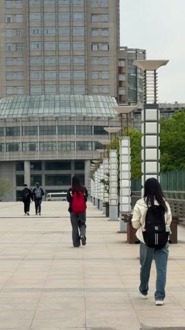 The video shows an outdoor scene in what appears to be a city or university campus, with several people walking in different directions on a paved plaza. In the foreground and midground, there are young adults with backpacks. One is wearing a white jacket and jeans with a black backpack, another has a red backpack and grey pants. A third person has a khaki-colored backpack. Two individuals in the distance walk away from the camera. Another man runs in the background. Modern architecture is visible in the background, including a large building with many windows. There are evenly spaced, decorative light posts along the plaza's edge. The weather is overcast.