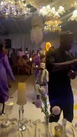 The video captures an indoor event, seemingly a wedding reception or party, based on the decorations and atmosphere. The scene is brightly lit by elaborate chandeliers hanging from a white-draped ceiling. The floor is reflective and features a large, stylized letter. A long table adorned with candles and floral arrangements sits in the foreground. Several people are dancing and moving around the dance floor, including some dressed in purple robes. Notably, a person in a gorilla costume is also dancing. People filming the event are also present, including one person with a camera visible in the center of the frame. The general atmosphere is lively and festive.
