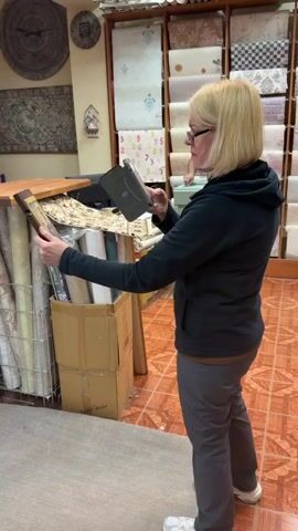 A woman with blonde hair and glasses is in a shop that appears to sell wallpaper and religious icons. She is holding a framed icon and a dark-colored device, possibly a phone or small camera, which she is using to take a picture of the icon. She is looking at the camera and smiling slightly. Behind her, shelves are filled with rolls of wallpaper. A computer monitor and other items are visible in the background. The flooring is tiled in a reddish-brown color. The overall lighting suggests it is daytime. The scene is likely in Maloyaroslavets, Russia, given the provided location context. The woman's action of photographing the icon suggests she is documenting it, perhaps for a customer or for her own records.