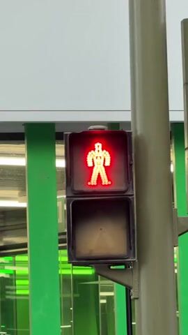 The video shows a pedestrian traffic light with a red silhouette of a person illuminated. The traffic light is attached to a pole on the right side of the frame. Behind the traffic light, a building with a white and green facade is visible. The lower part of the building has large glass windows and green vertical supports. The video pans slightly to reveal a street and sidewalk in front of the building, with a tree on the left. Two people are visible on the sidewalk. One person is walking away from the camera on the left side of the sidewalk, and another person is walking towards the camera on the right side of the sidewalk. The traffic light remains red throughout the short clip.
